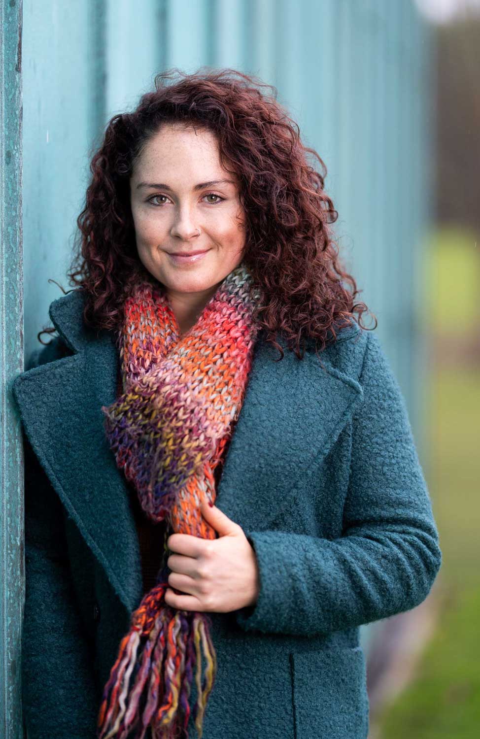 Woman in a teal coat and colourful knitted scarf leaning against a painted wall in Newport