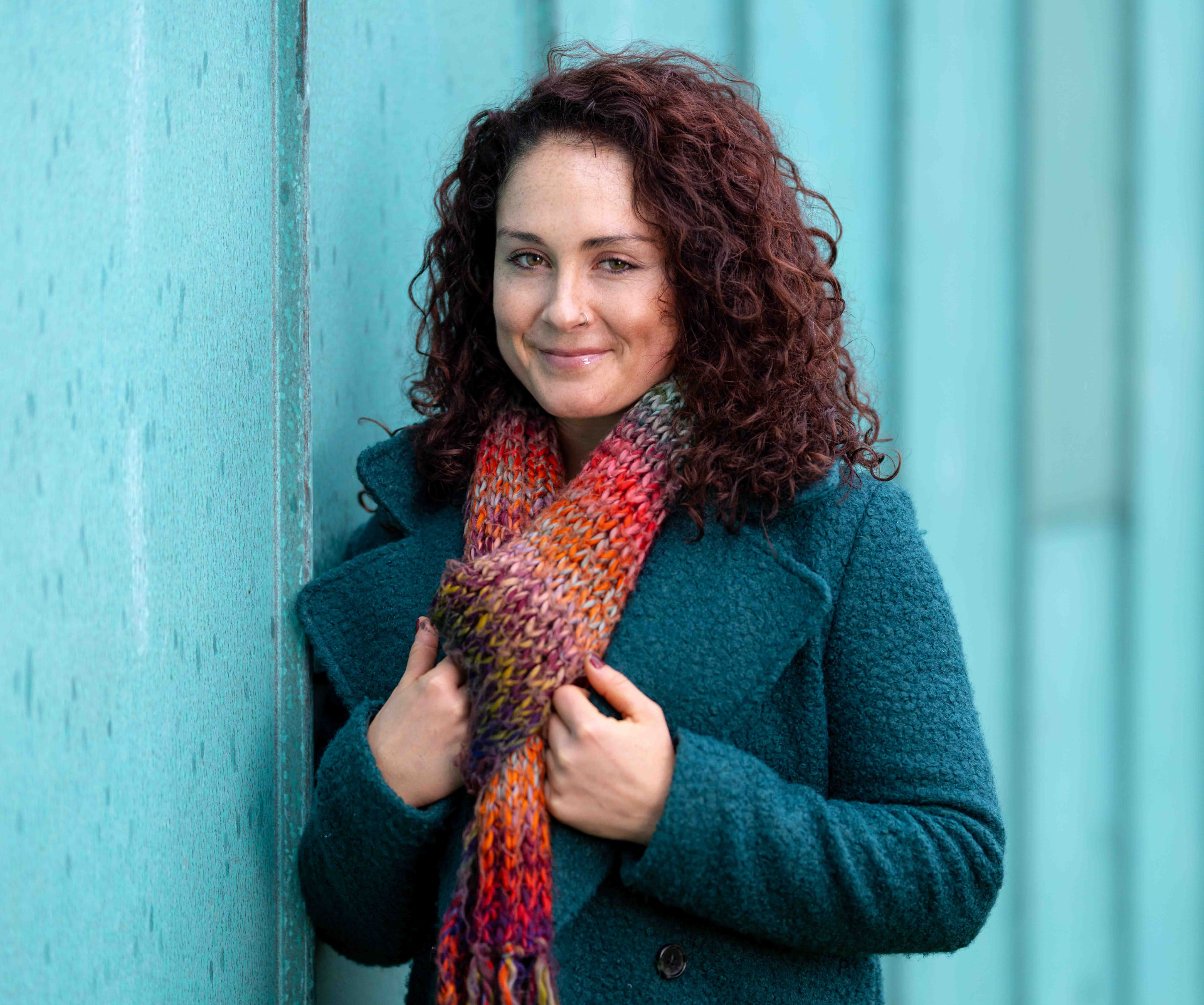 Portrait of a woman in a teal coat and knitted scarf against a turquoise wall in Newport