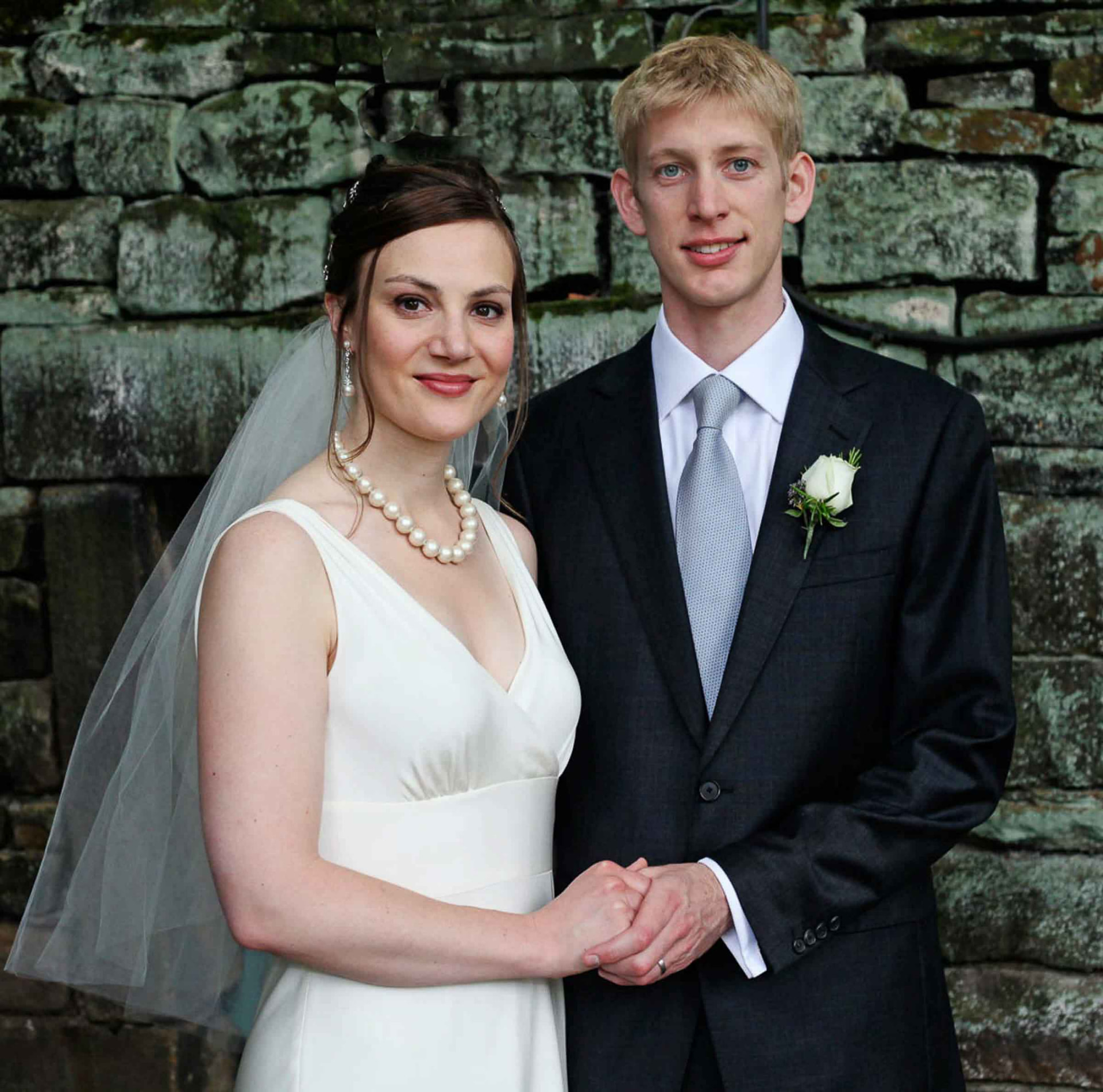 Newlyweds holding hands in front of a moss-covered stone wall, Welsh countryside wedding