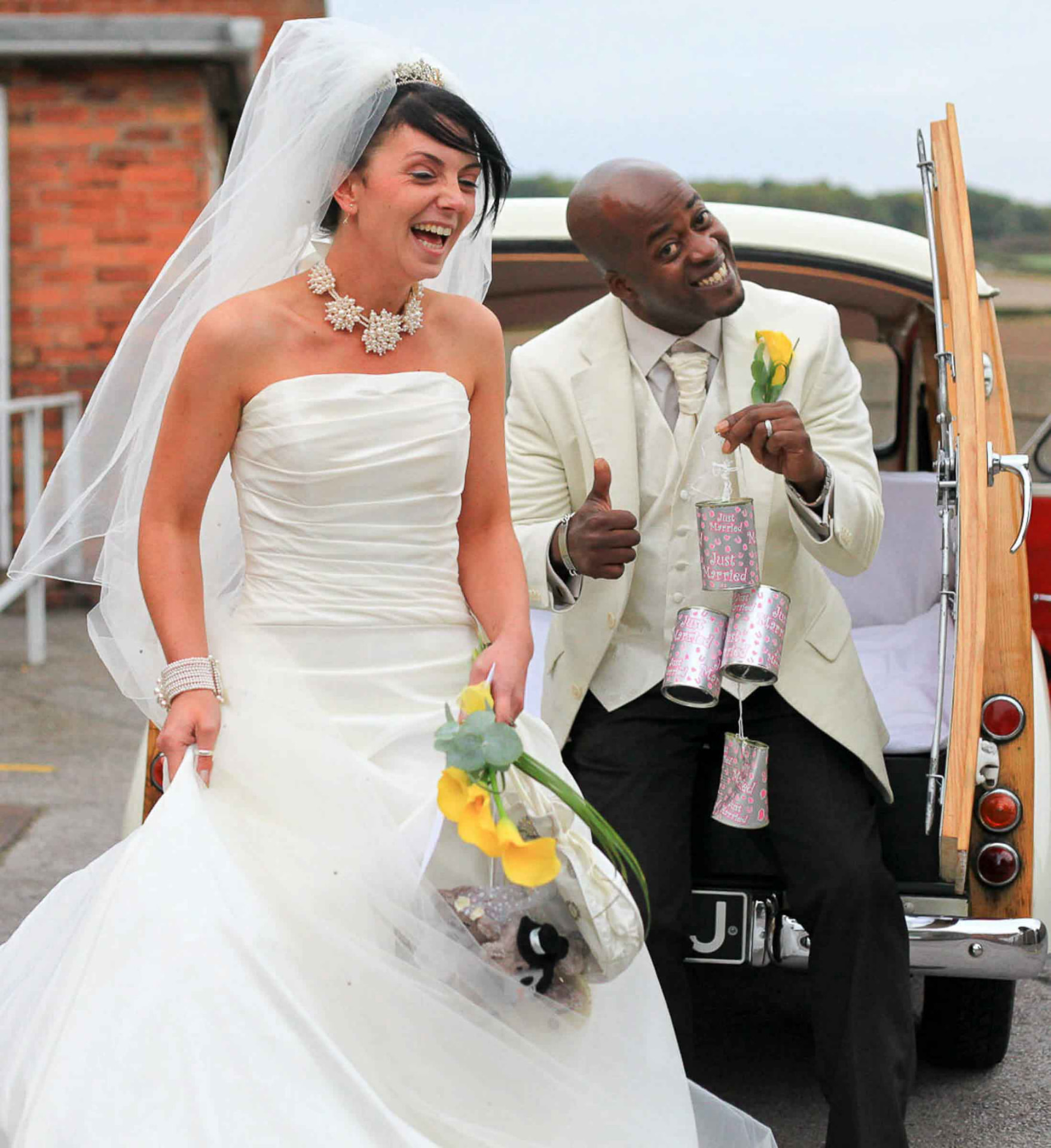 Bride and groom laughing beside a vintage wedding car decorated with just-married tins, South Wales
